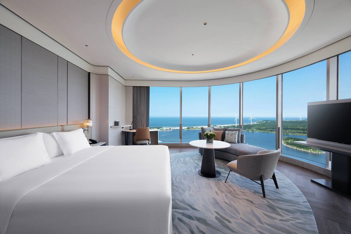 A modern hotel room with a large bed, a sofa, a small table, and a TV. The room has large windows offering a view of the ocean and wind turbines.