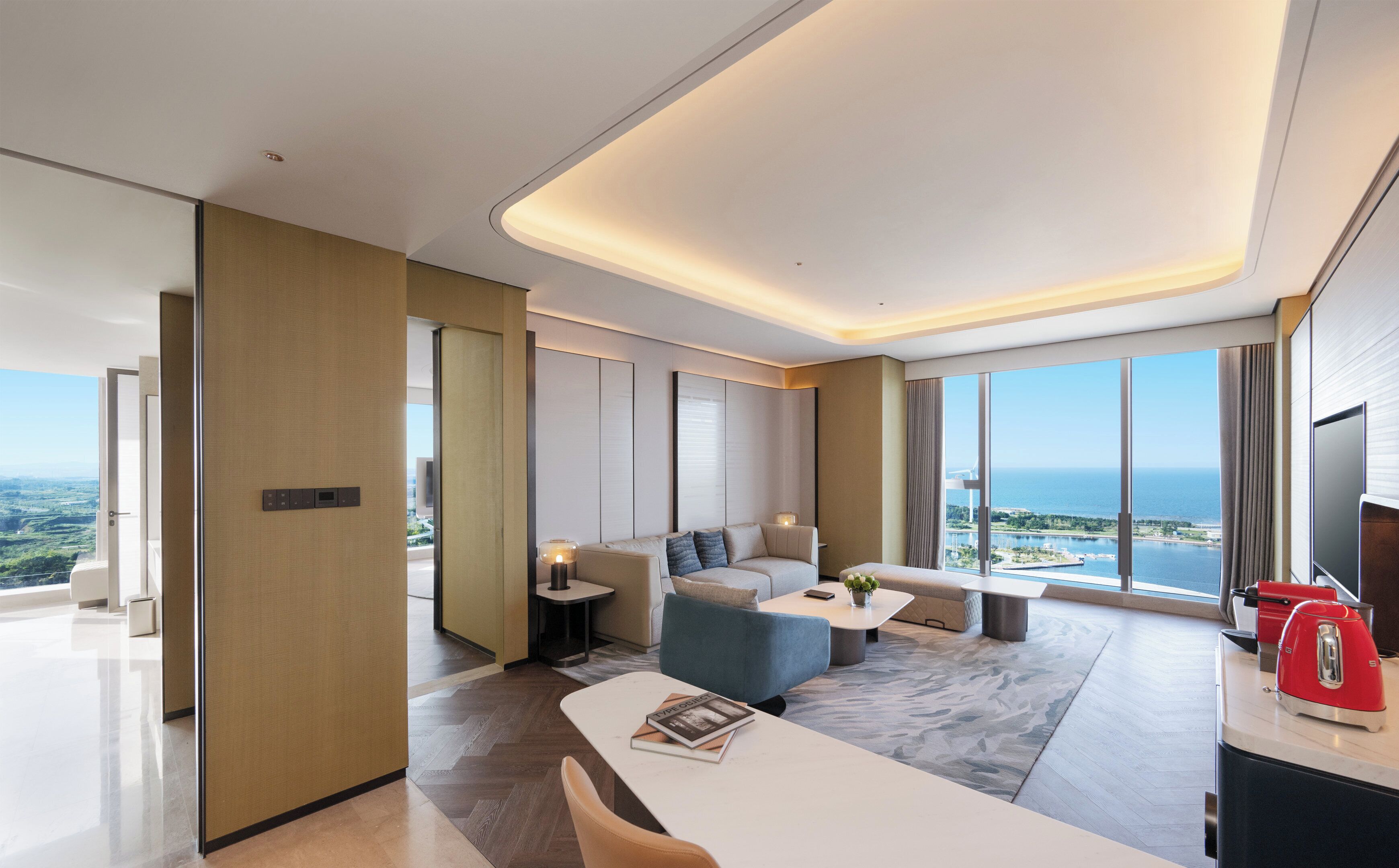 Habitación de hotel moderna con un ventanal que ofrece vista al mar, un sofá, una mesa de centro y un comedor.