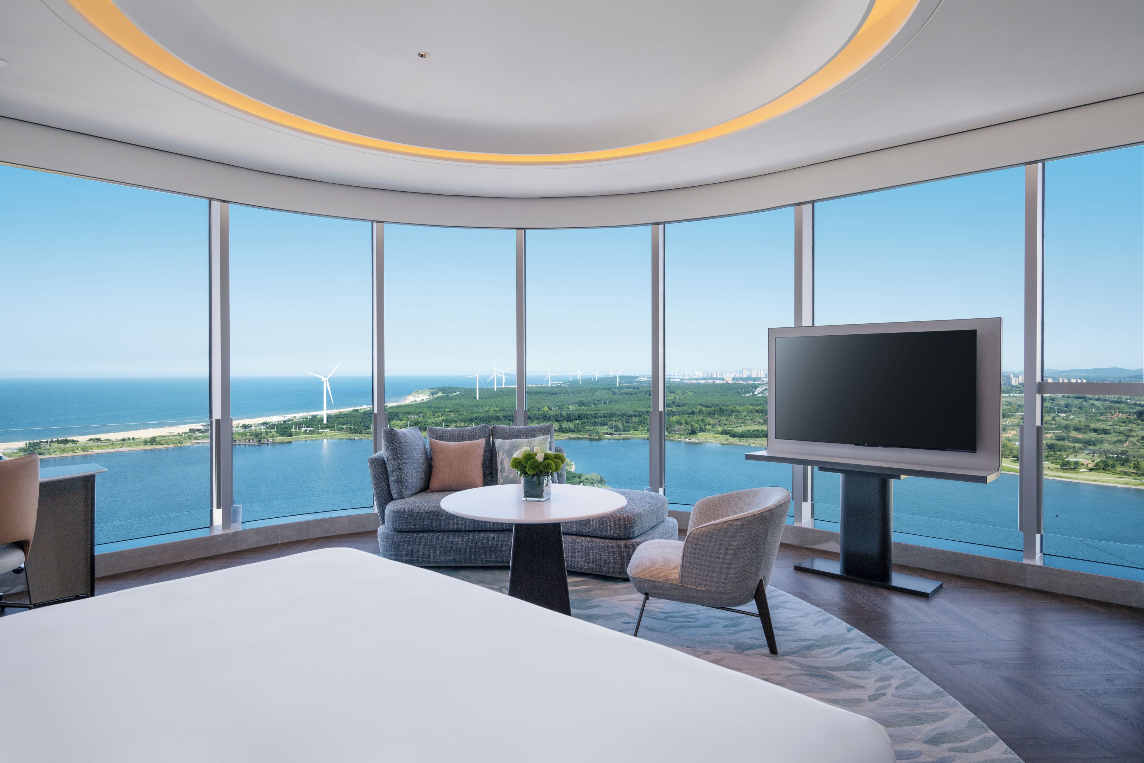 A modern hotel room with a large bed, a sofa, a TV, and a view of the sea and wind turbines.
