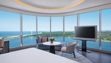 A modern hotel room with a large bed, a sofa, a TV, and a view of the sea and wind turbines.