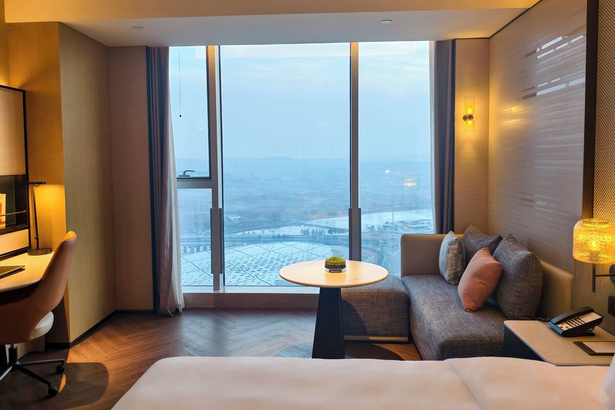 A hotel room with a large window, a bed, a sofa, a desk, and a chair.