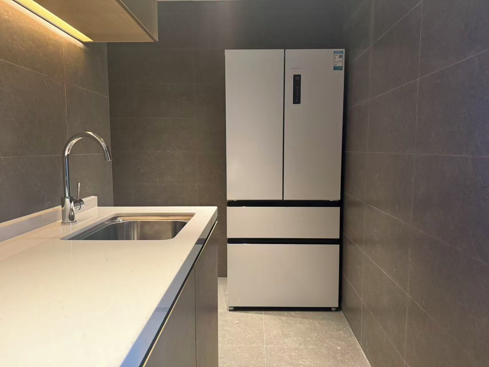 A modern kitchen with a stainless steel sink, a built-in refrigerator, and dark tiled walls.