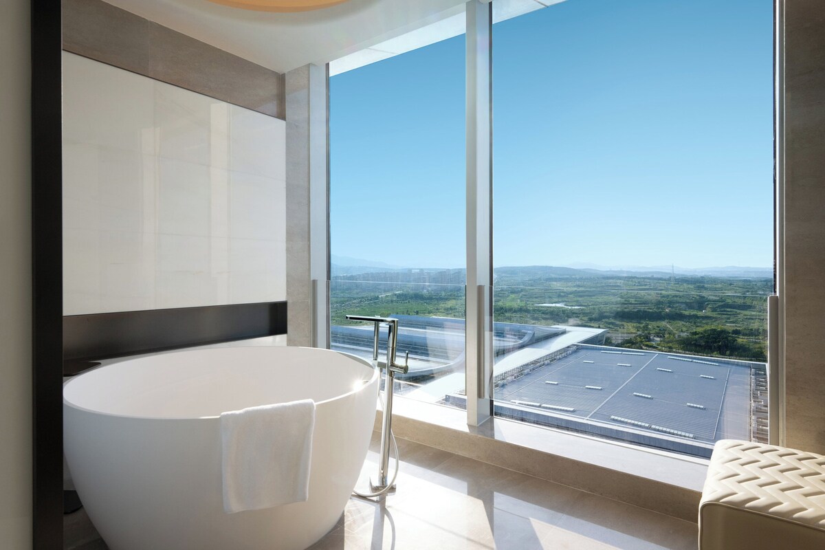 A modern bathroom with a large bathtub, a glass shower enclosure, and a view of a cityscape through large windows.