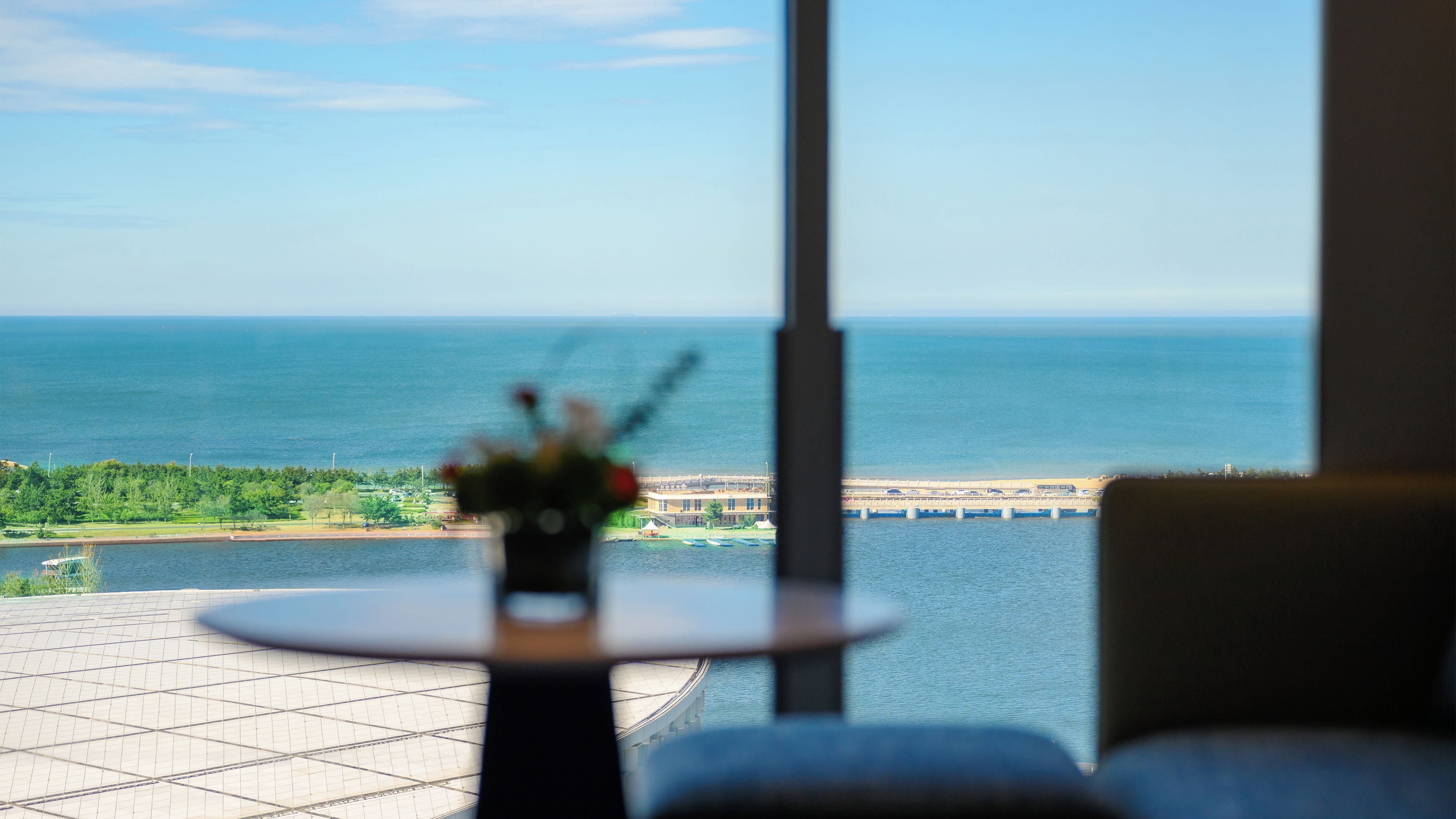 A view of a large body of water, a bridge, and a distant shoreline through a window with a small table and chairs in the foreground.