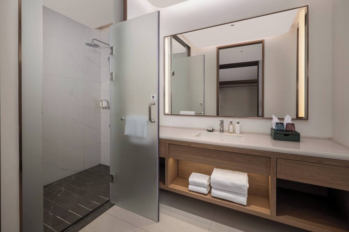 Un baño moderno con puerta de ducha de vidrio, un espejo grande y un mueble de tocador de madera con lavamanos y espacio para toallas.