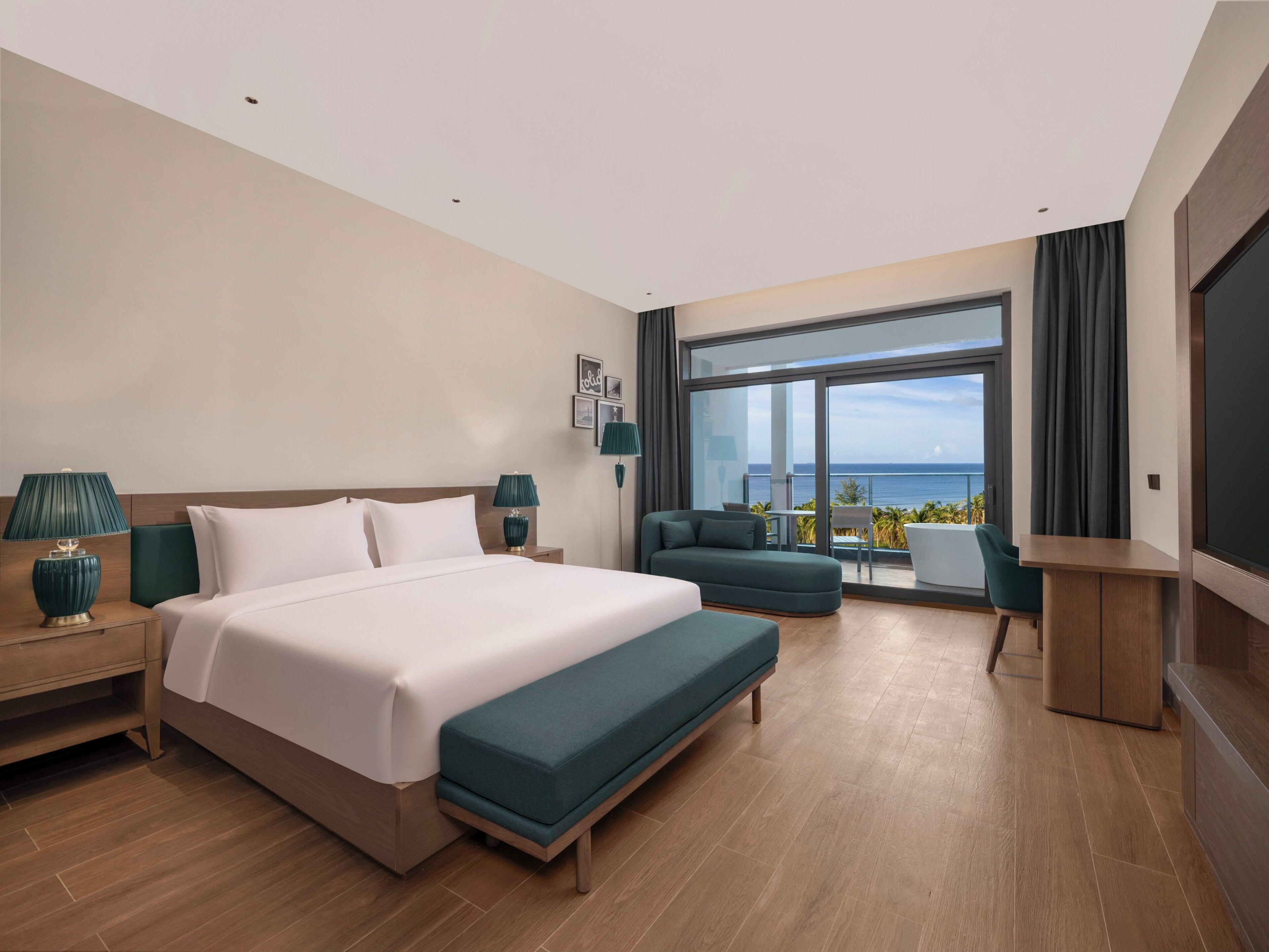 A hotel room with a large bed, a sofa, a small table, and a TV. There is a balcony with a view of the ocean.