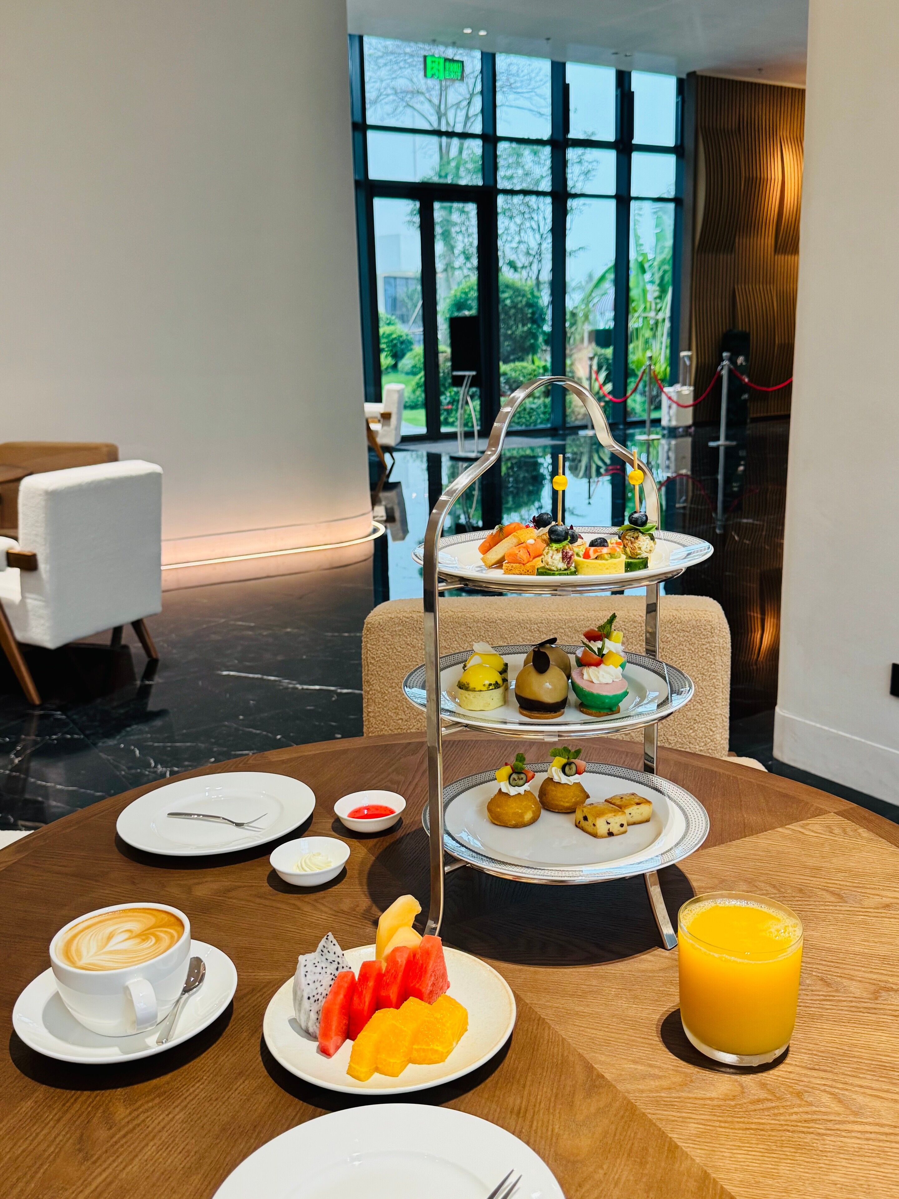 A dining table with a three-tiered dessert stand, plates of fruit, a cup of coffee, and a glass of orange juice.