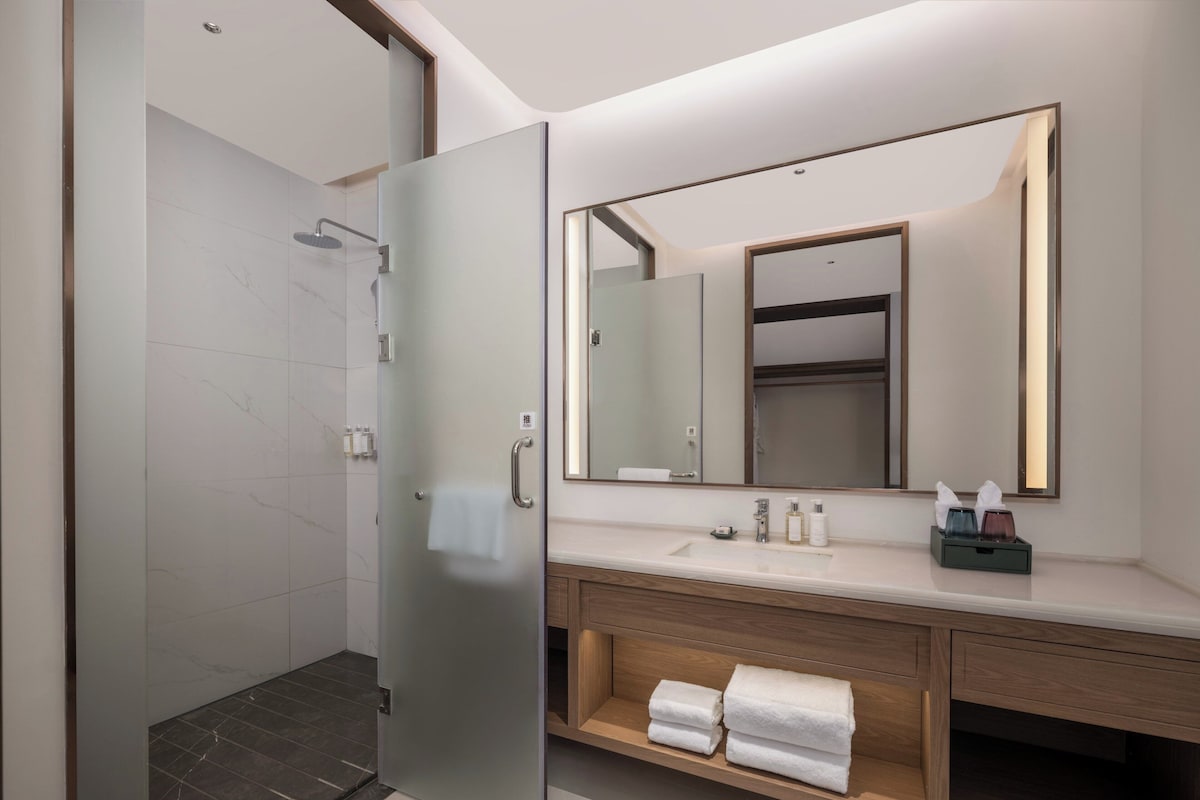 Un baño moderno con puerta de ducha de vidrio, un espejo grande y un mueble de tocador de madera con lavamanos y espacio para toallas.