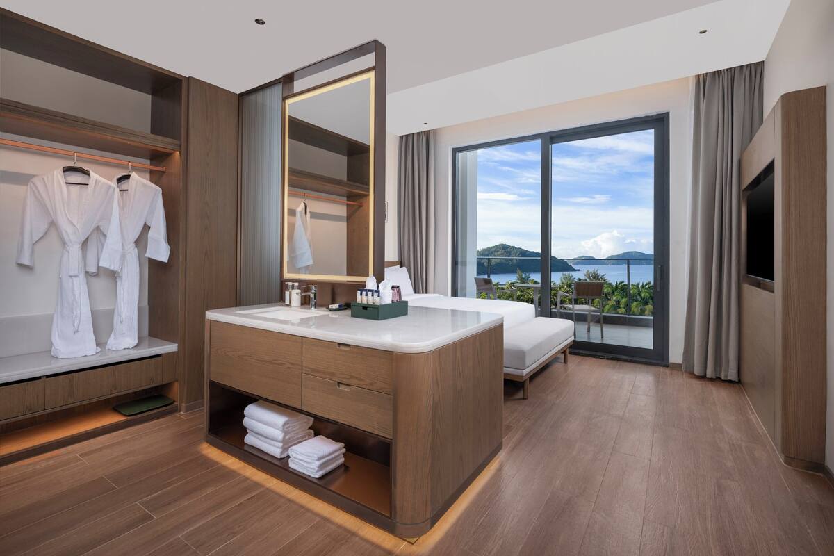 Habitación de hotel moderna con una cama grande, un tocador con espejo, un baño con ventanal grande y vistas al mar.