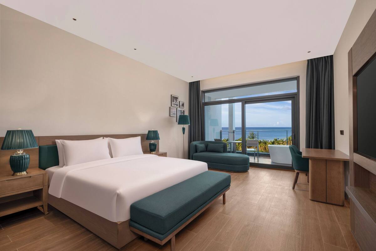 Habitación de hotel con una cama grande, un sofá, una mesita y un televisor. Hay un balcón con vista al océano.