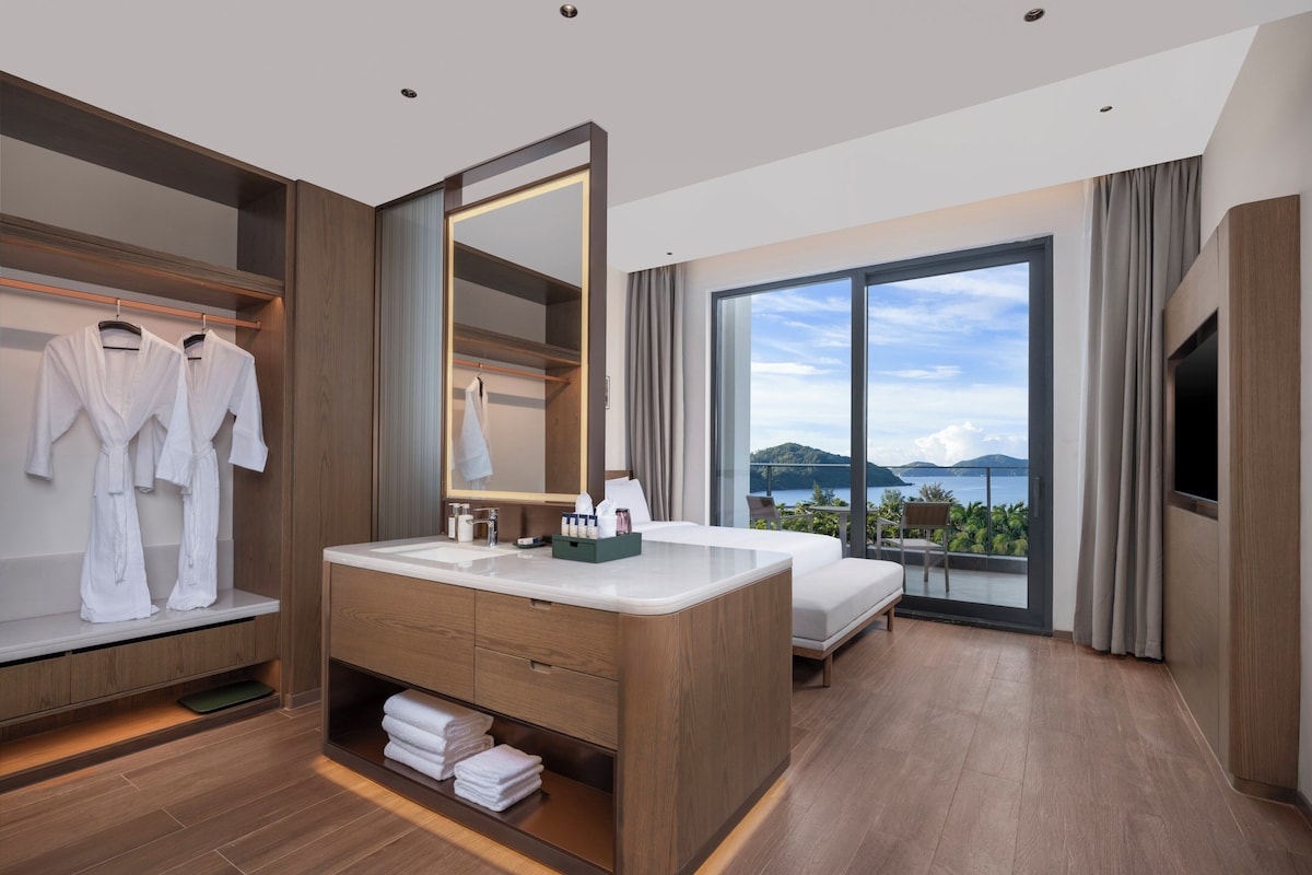 Habitación de hotel moderna con una cama grande, un tocador con espejo, un baño con ventanal grande y vistas al mar.