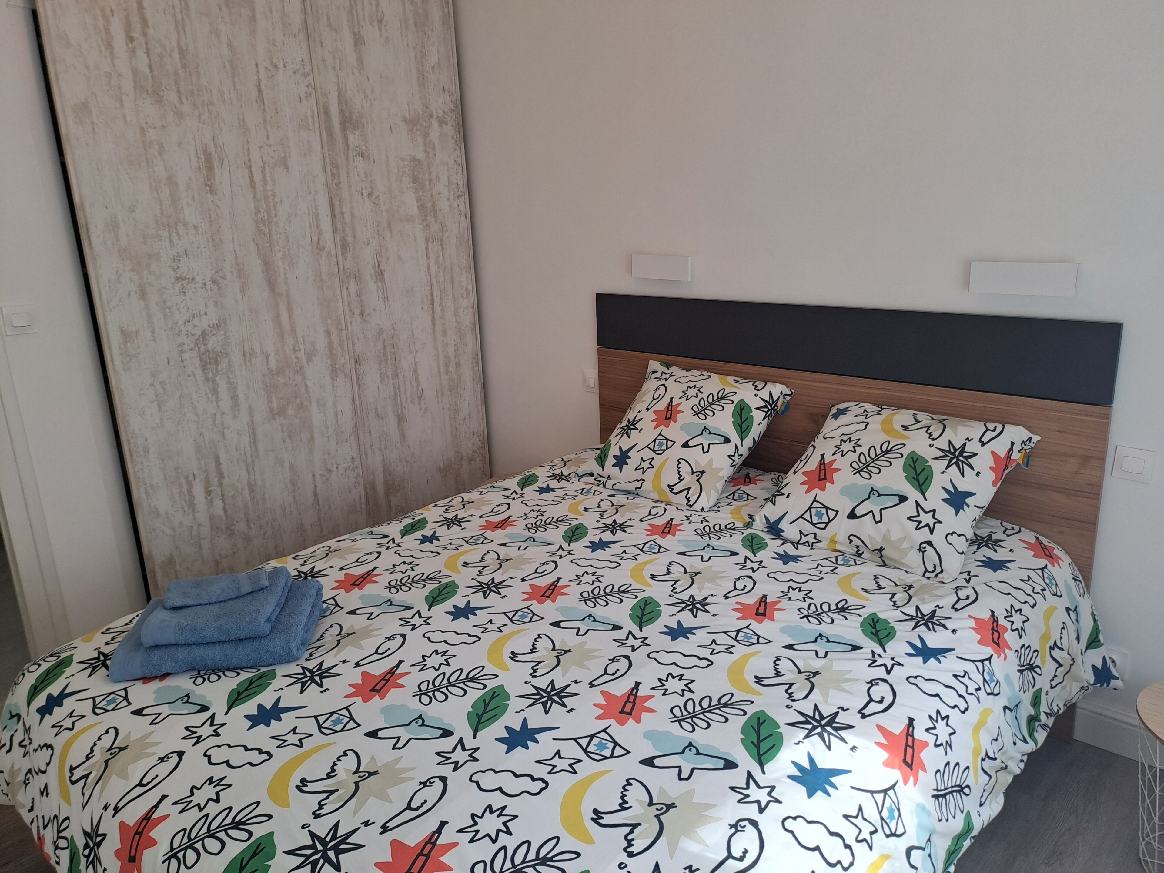 Un llit amb un estovat estampat i ple de color, una tovallola blava ben arrugada i un capçal de fusta.