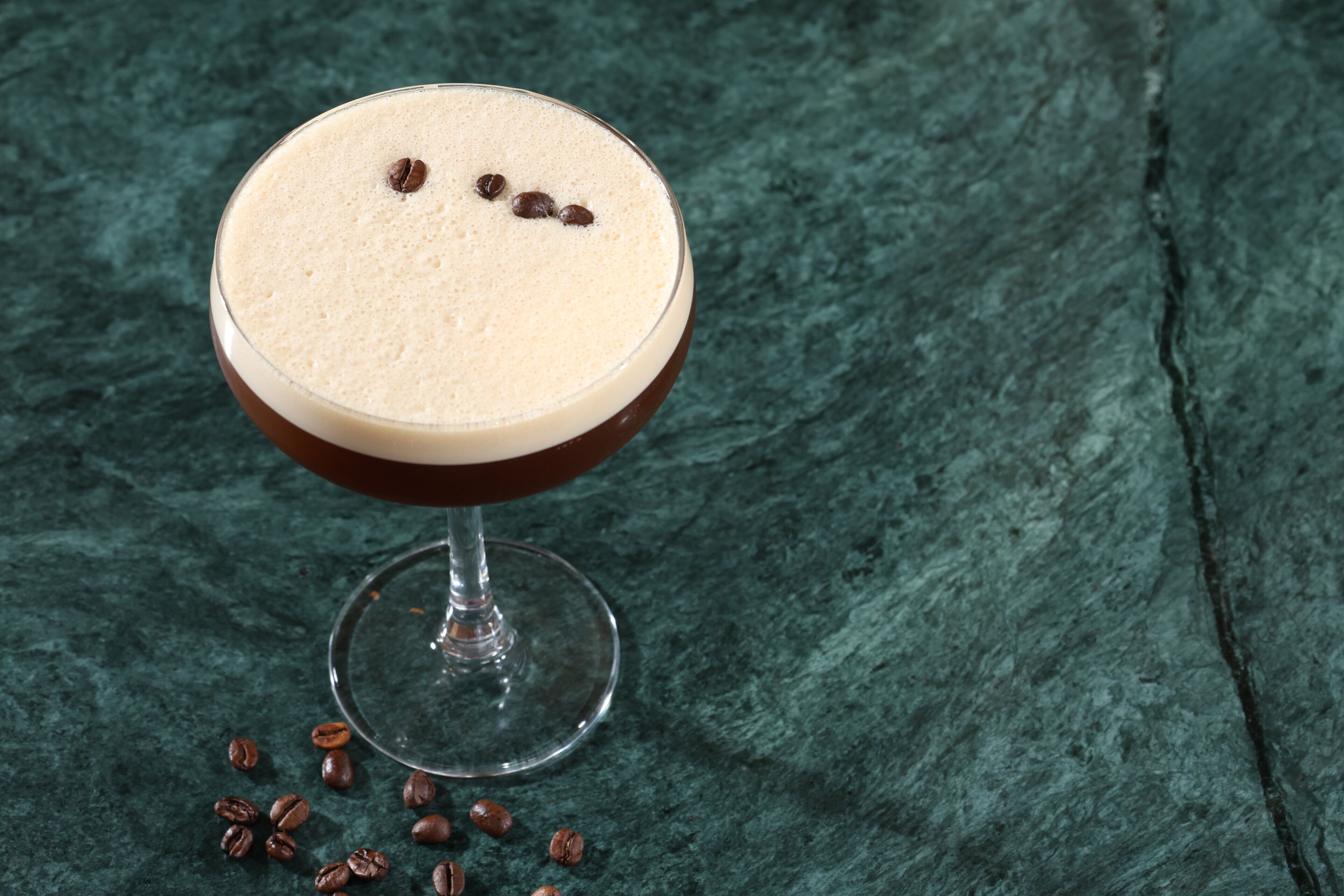 A cocktail glass with a frothy top and coffee beans on a textured surface.
