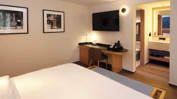 Habitación de hotel con cama, escritorio, silla, televisión de pantalla plana, cuadros en la pared y baño visible a través de una puerta abierta.