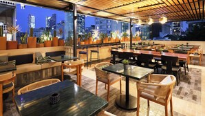 A rooftop restaurant with outdoor seating, city views, and potted plants.