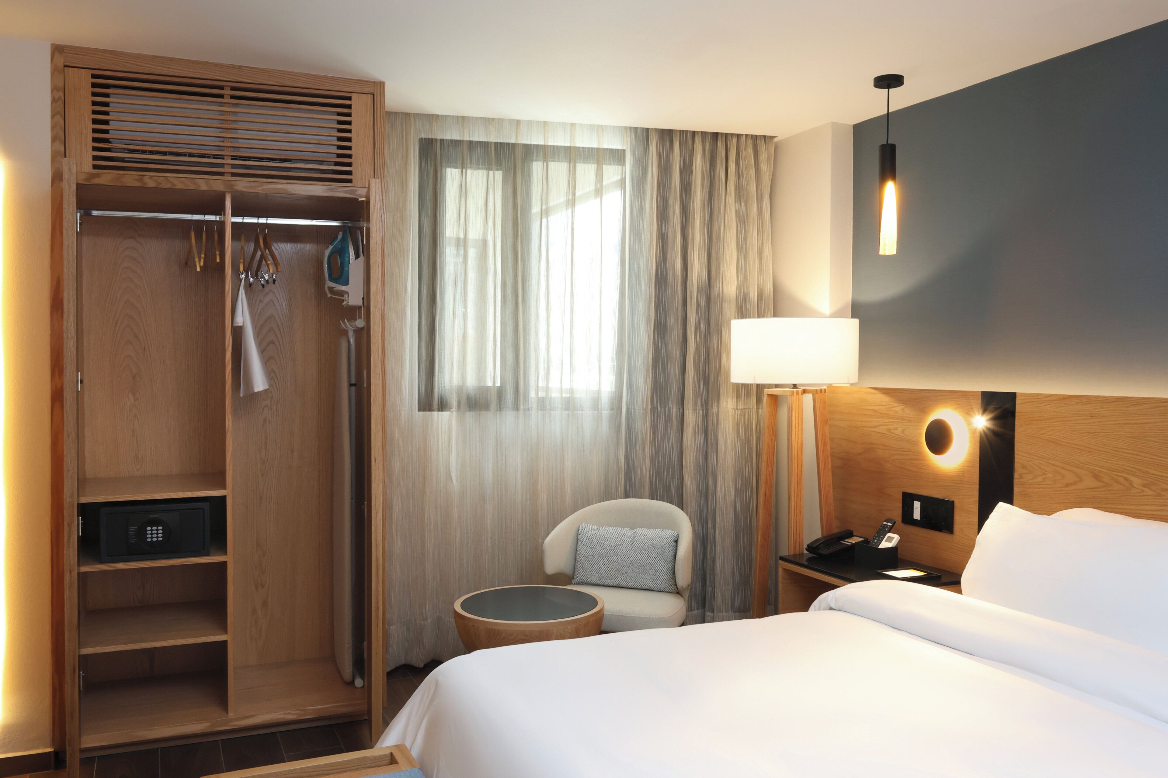 Et hotelværelse med seng, stol, natbord, lampe, garderobeskab og et vindue med gardiner.