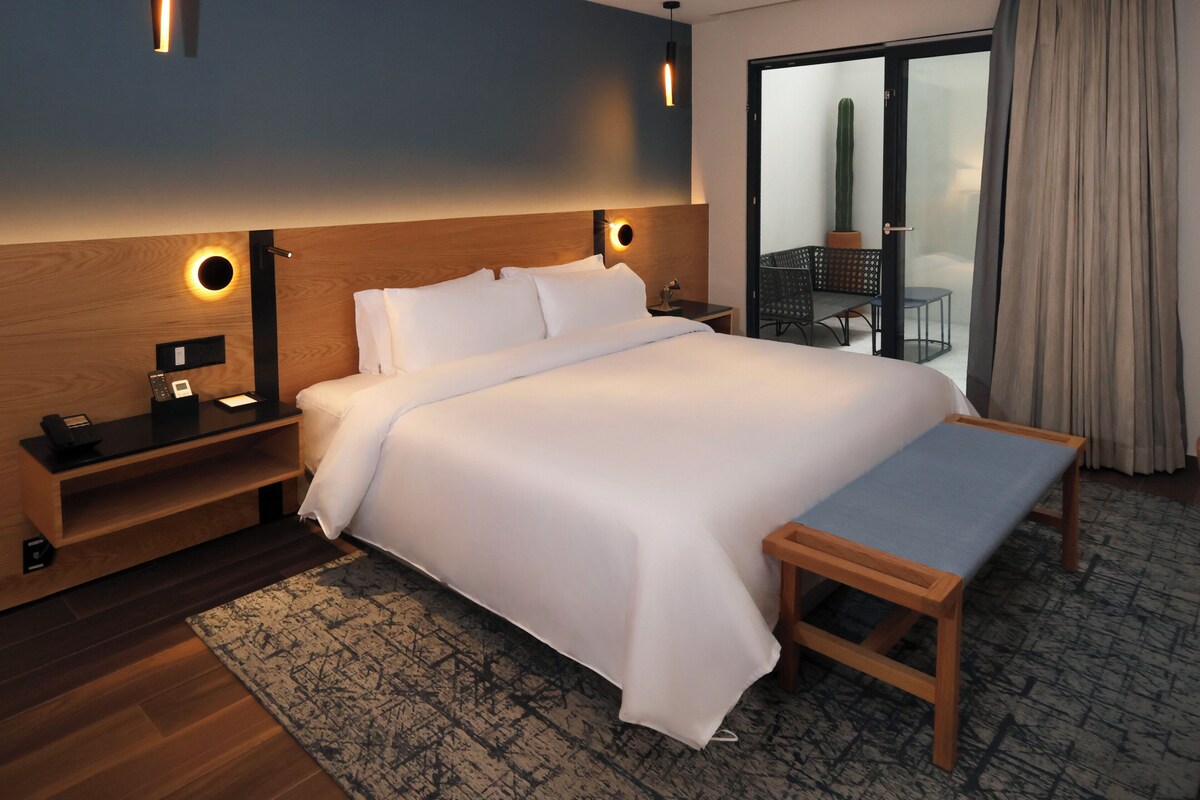 Habitación de hotel con una cama grande, mesitas de noche, un banco, una pequeña mesa y vistas a un cactus a través de una puerta corrediza de vidrio.