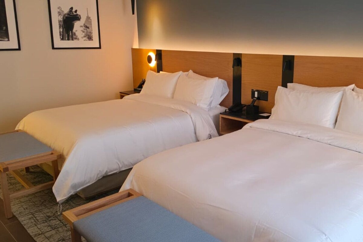 Habitación de hotel con dos camas, una mesita de noche, una lámpara y un cuadro en la pared.