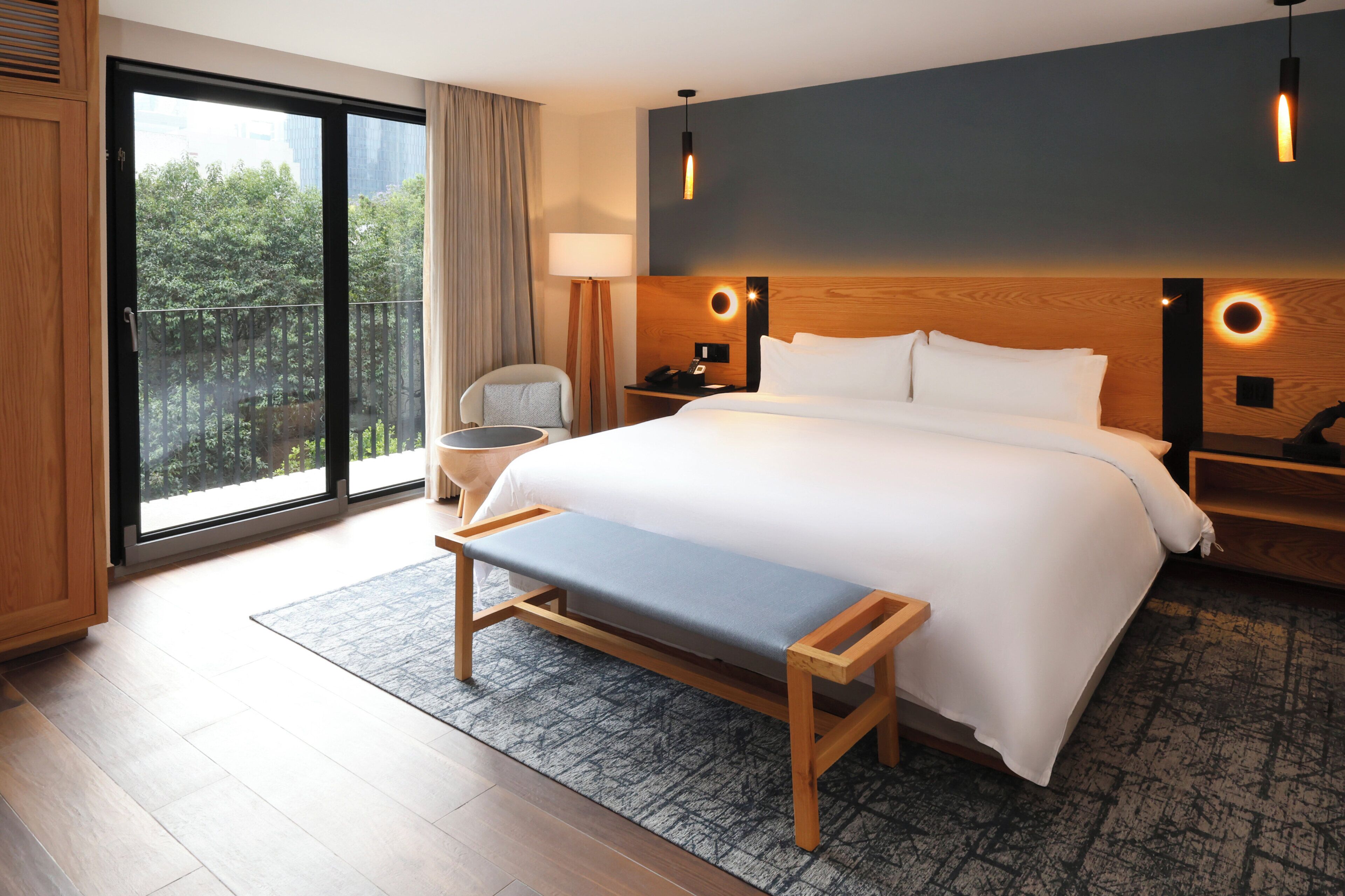 A hotel room with a large bed, a bench, a desk, a chair, a nightstand, a lamp, and a view of greenery through the window.