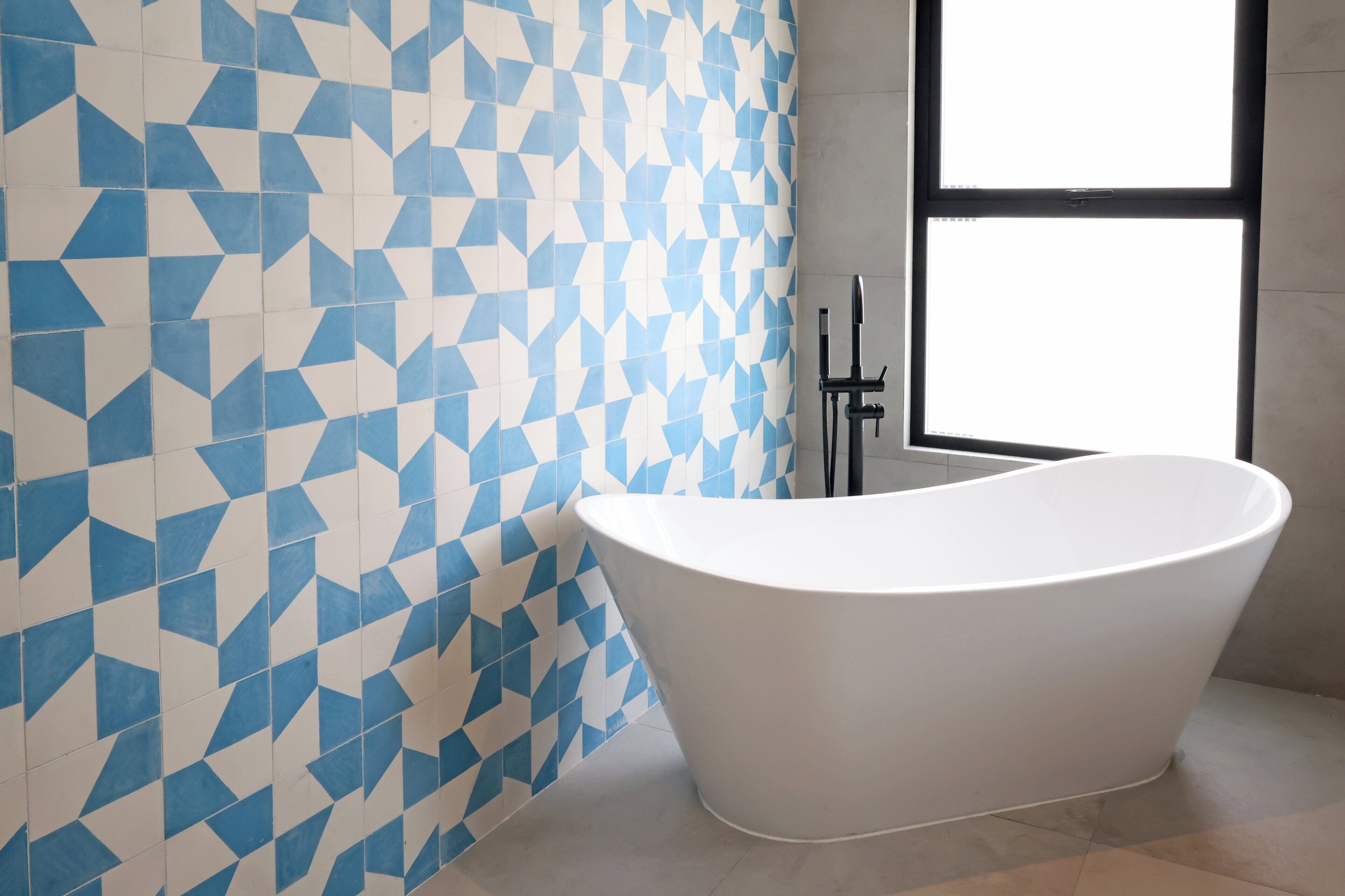 A modern bathroom with a geometric tile wall, a freestanding bathtub, and a window providing natural light.