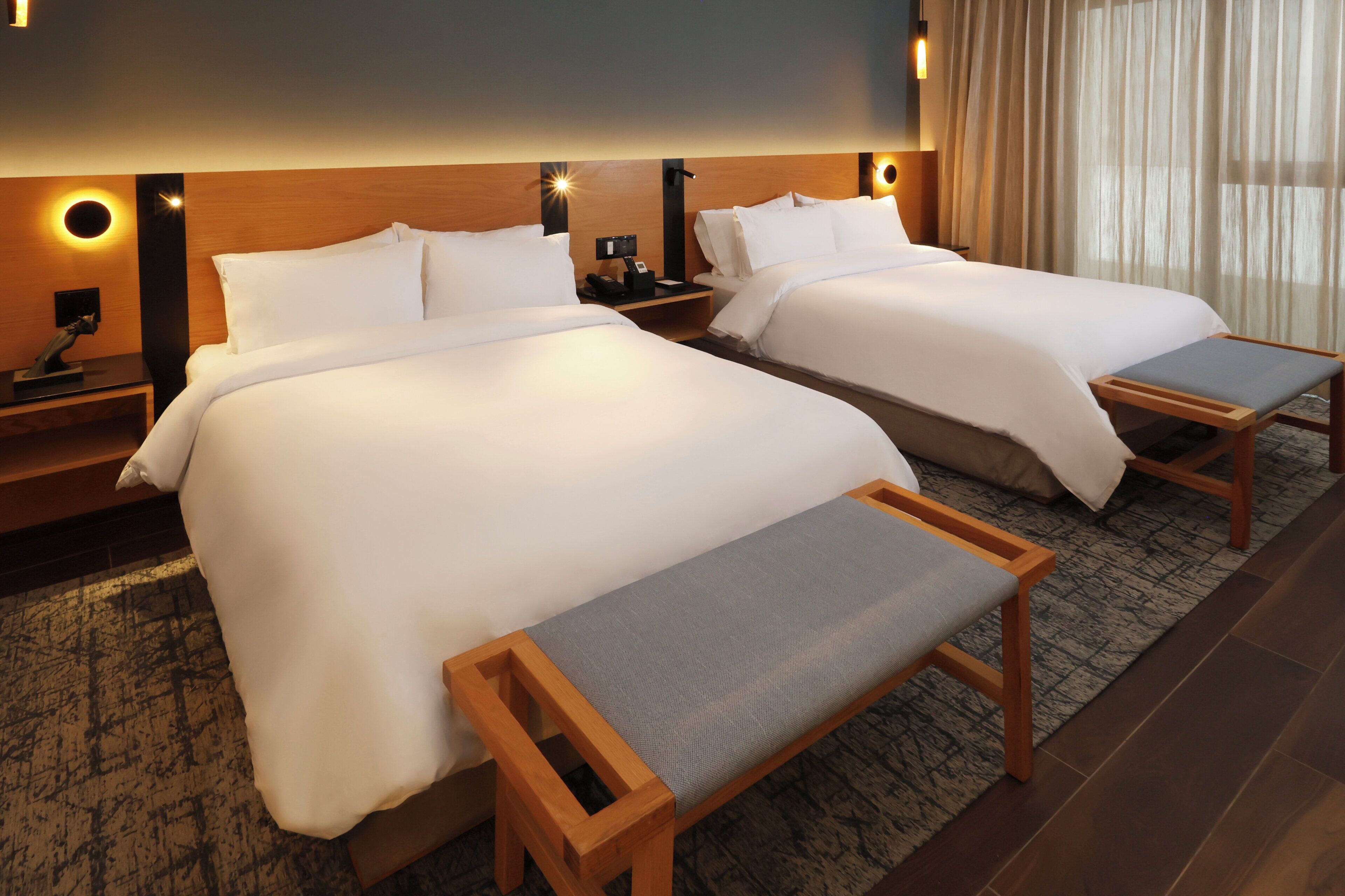 A hotel room with two beds, bedside tables, a bench, and a large window with curtains.