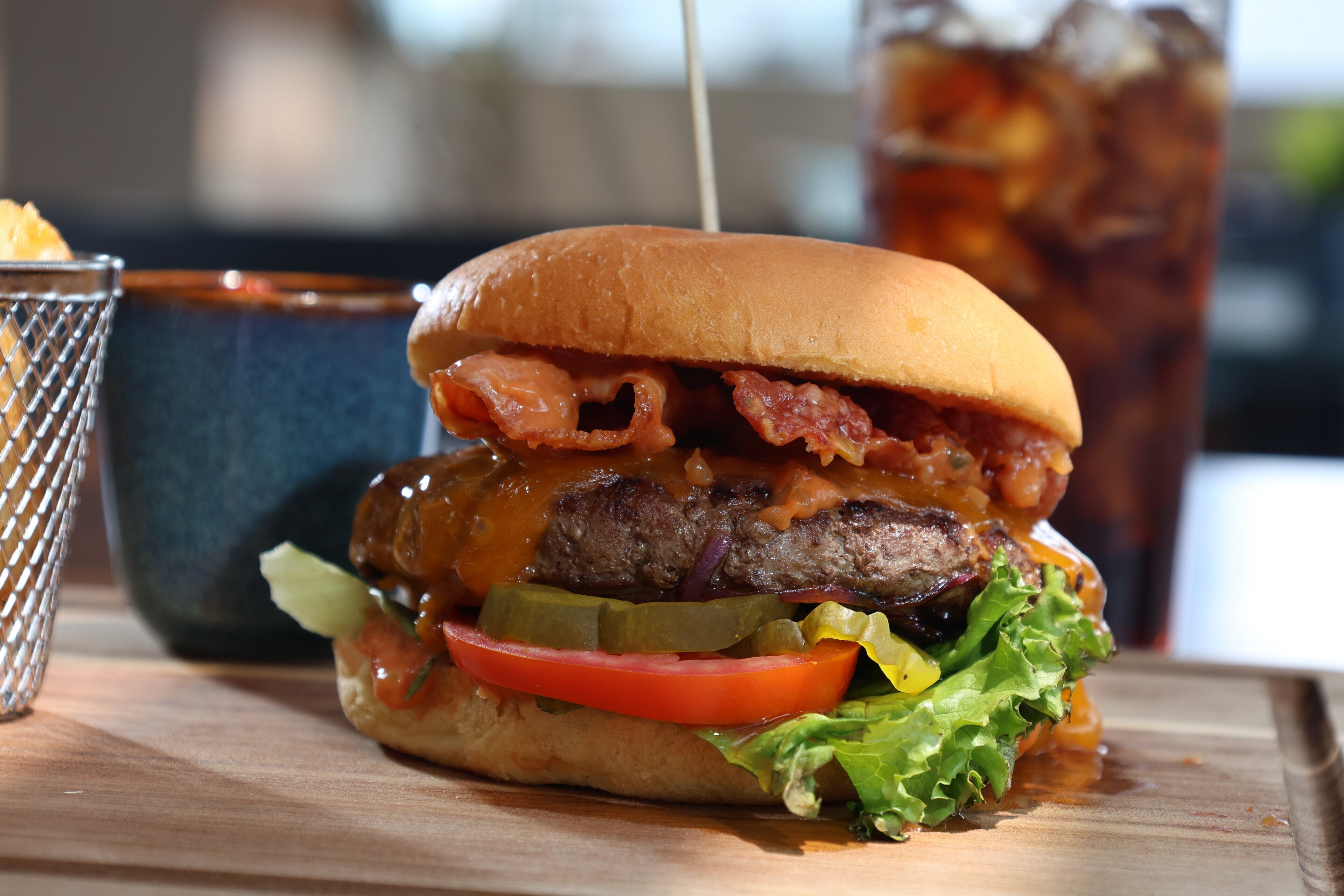 A burger with bacon, cheese, and lettuce on a bun, with a drink in the background.