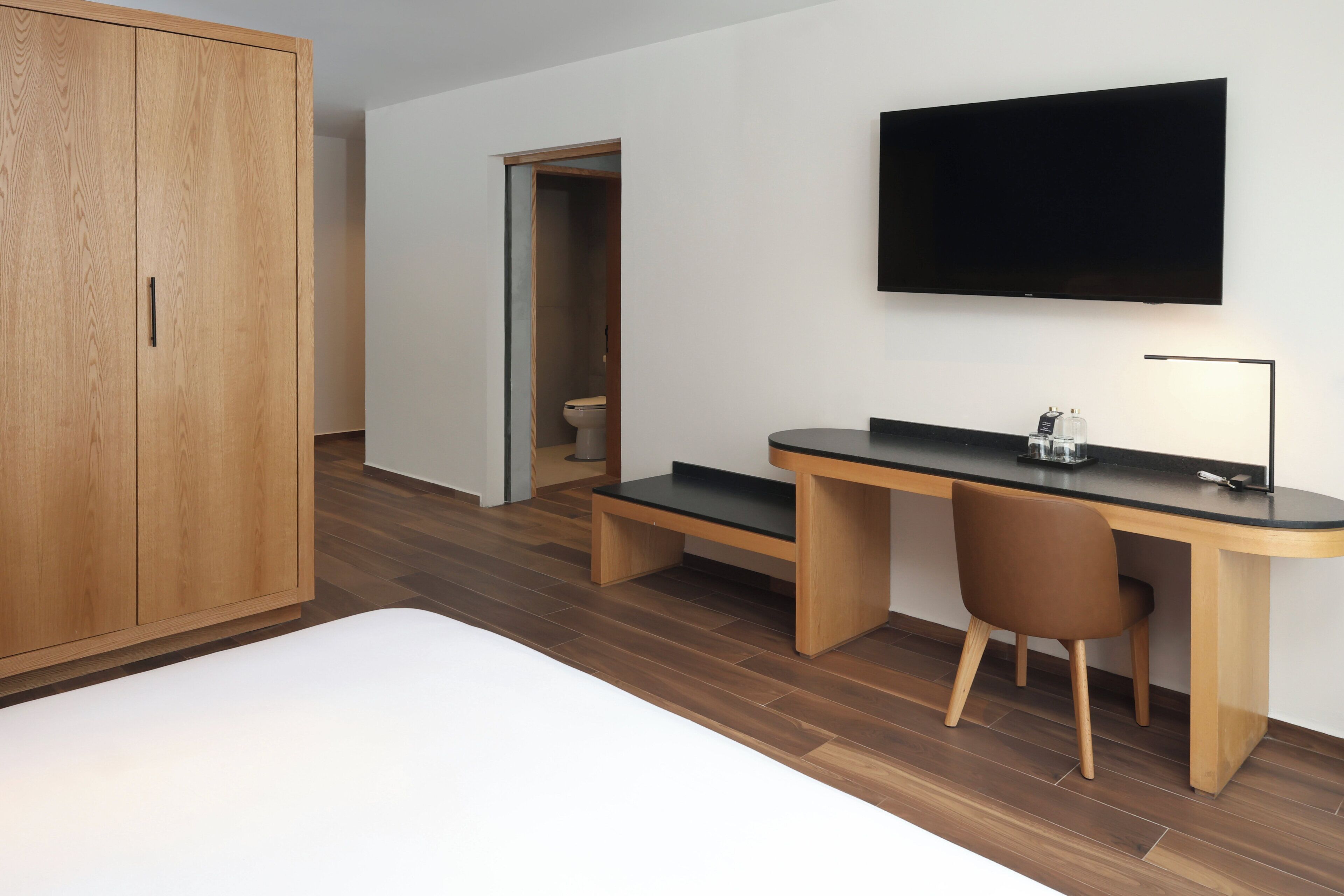 Un dormitorio moderno con un armario de madera, un escritorio con una silla, un televisor y un inodoro visible a través de una puerta abierta.