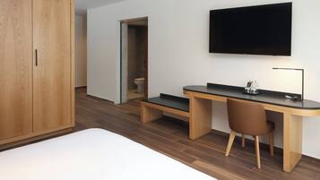 Un dormitorio moderno con un armario de madera, un escritorio con una silla, un televisor y un inodoro visible a través de una puerta abierta.