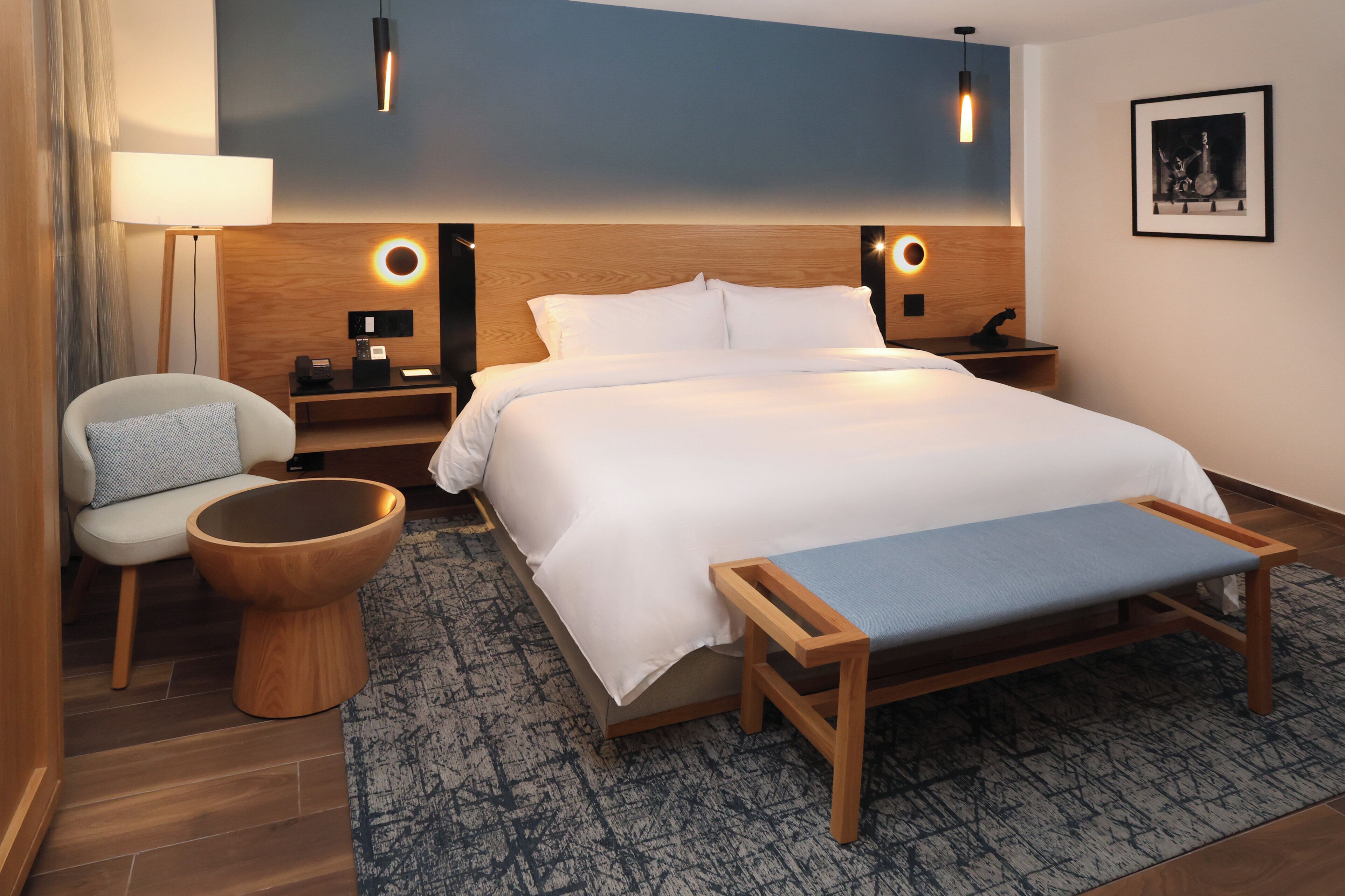 Une chambre d’hôtel moderne avec un grand lit, des tables de chevet, une chaise, un tabouret rond en bois et un banc.