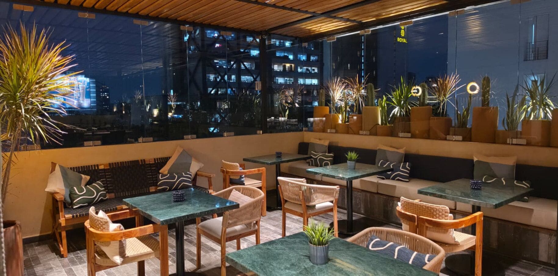An outdoor seating area with tables and chairs, potted plants, and a view of a cityscape at night.