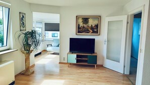 Un salon moderne avec un téléviseur à écran plat posé sur un meuble en bois, une grande plante en pot et un tableau accroché au mur.