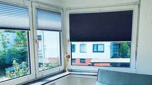 Une pièce compacte dotée d’un bureau intégré, d’un canapé gris et de grandes fenêtres offrant une vue sur les bâtiments et la verdure.
