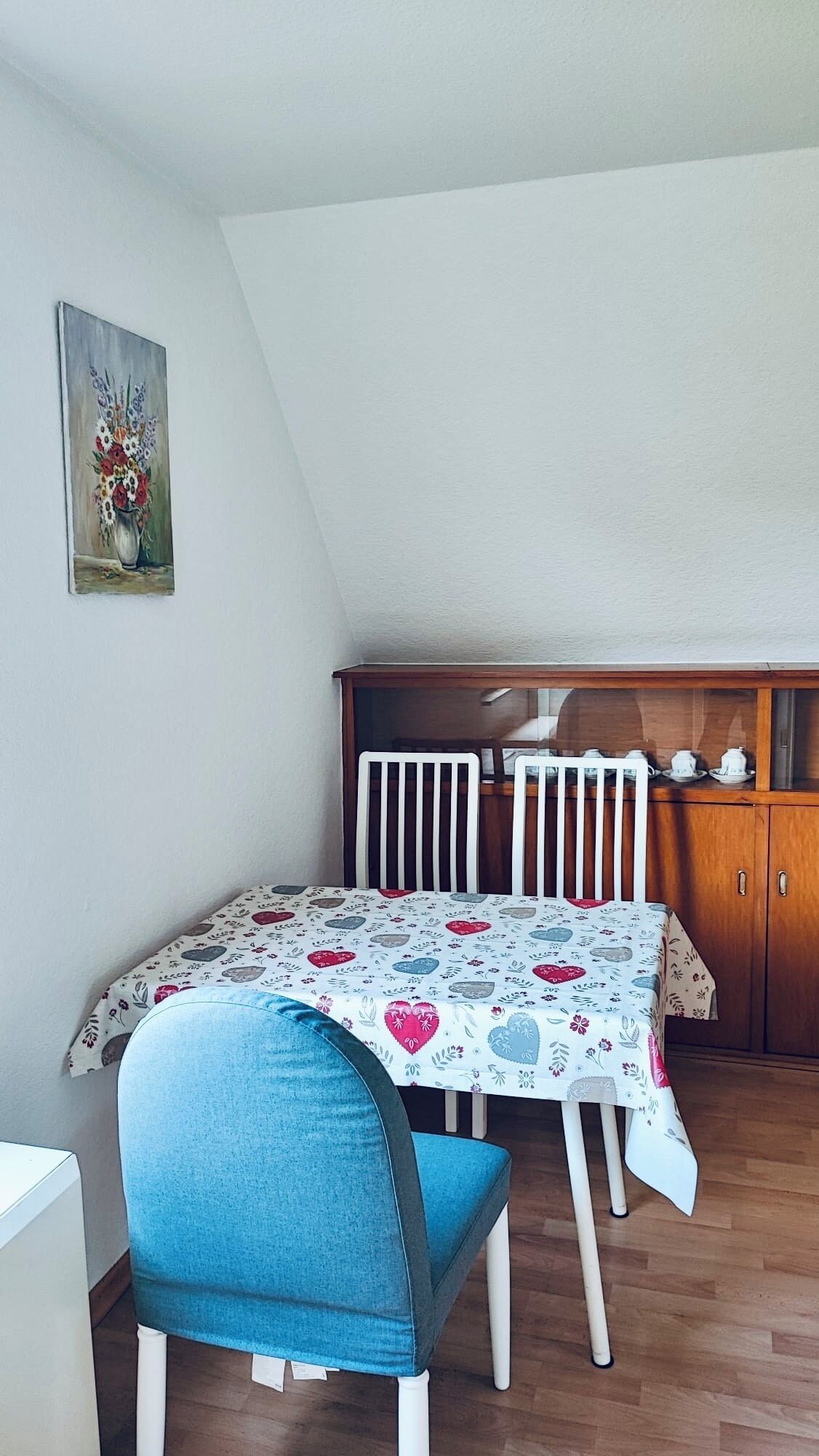 Ein Essbereich mit Tisch für zwei Personen, einem blauen Stuhl, einer Holzkabine mit Teeservice und einem Bild an der Wand.