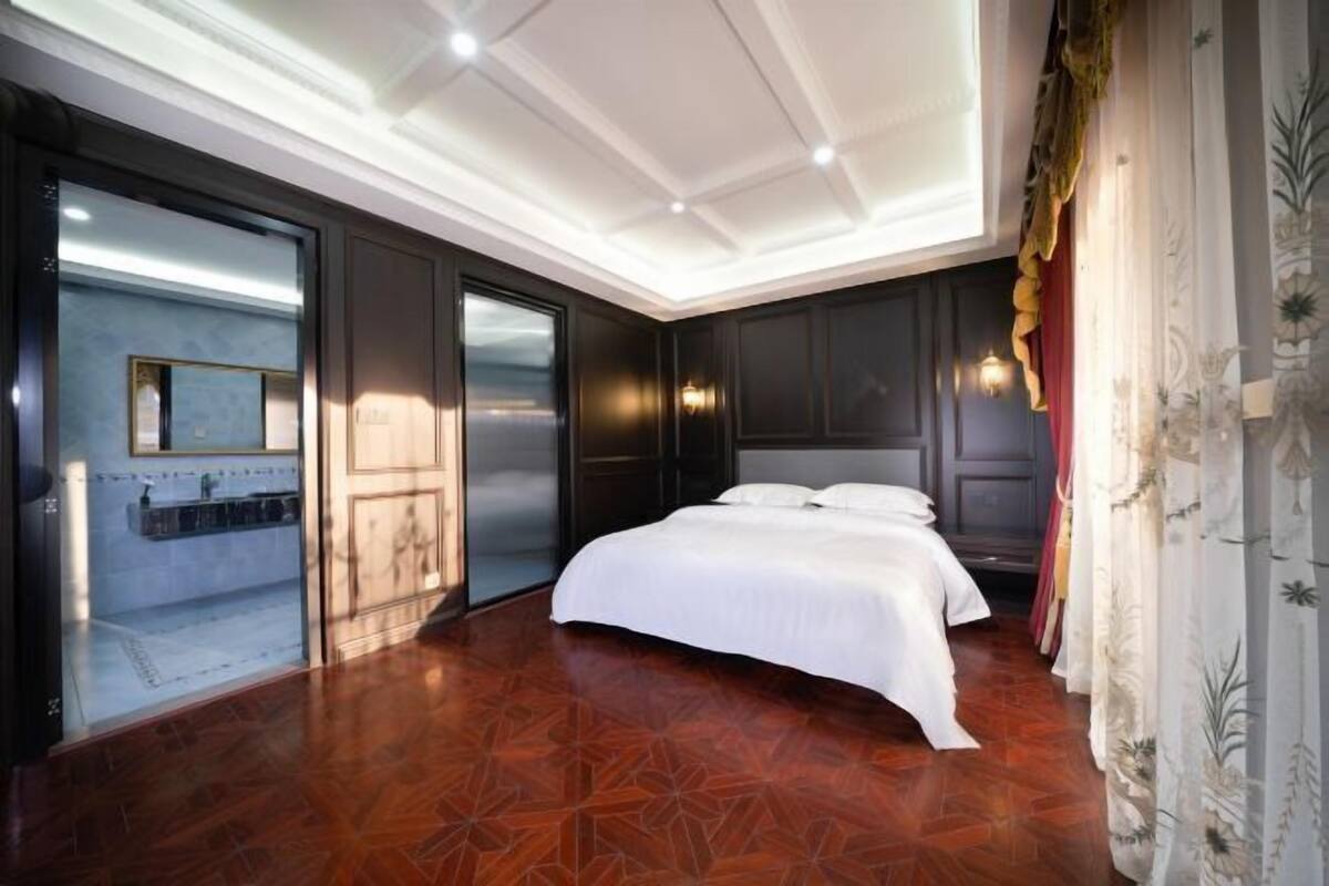 A bedroom with a large bed, a mirror, a wardrobe, and a bathroom visible through a glass door.