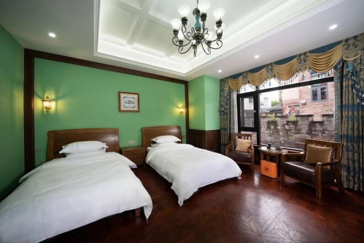 A hotel room with two beds, a chandelier, a window with curtains, and a view of the street.