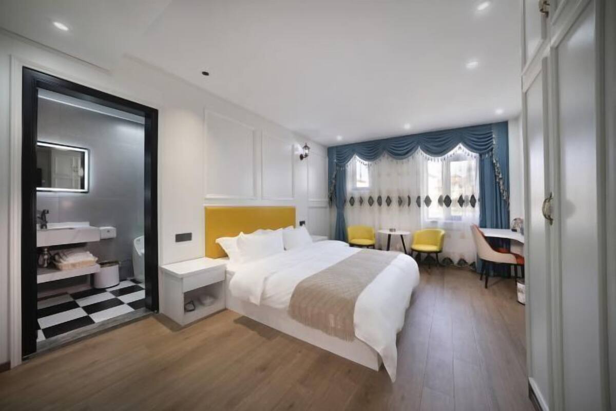 A modern hotel room with a large bed, a desk, a chair, a small table, and a bathroom with a checkered floor.