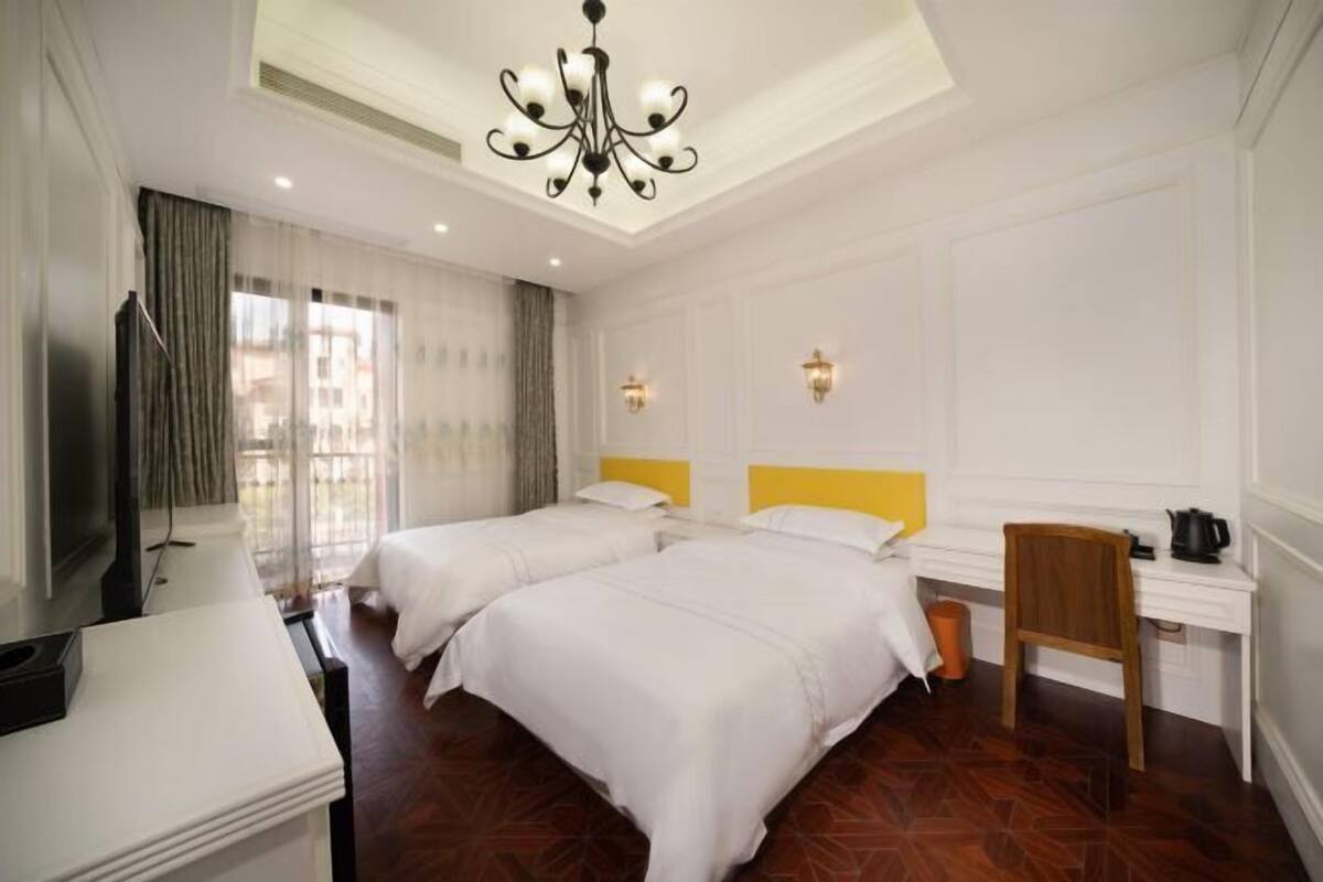 A hotel room with two beds, a desk, a chair, a TV, and a chandelier.