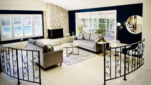 A living room with a stone fireplace, a sofa, a coffee table, and a dining area with a circular mirror. - Pacific Dreams at Woodland Hills Distinguished 8 Br 6 5 BA Residence With Pool and Spa Sleeps 16 (Calabasas)