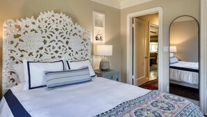 Habitación de hotel con una cama grande, un cabecero decorativo, una mesita de noche con lámpara, un espejo y vista al pasillo.