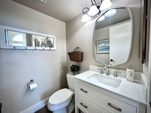 A modern bathroom with a toilet, sink, and mirror. There is a basket on the wall and a plant on the counter. - Crestview Oceanside - Family-friendly Private Yard (Oceanside)