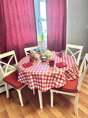 A dining table with a red and white checkered tablecloth, surrounded by white chairs with red cushions. A teapot, cups, and a bowl are on the table.
