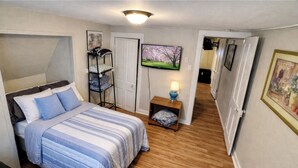 A bedroom with a bed, a nightstand with a lamp, a TV on the wall, and a door leading to another room.