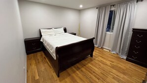 Ein Schlafzimmer mit Holzboden, einem Bett mit weißer Bettwäsche, einem Nachttisch, einer Kommode und einem Fenster mit Vorhängen.
