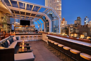 A rooftop bar with a glass enclosure, seating, bar stools, and a bar stocked with bottles.