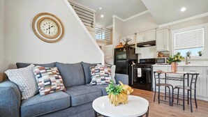 Una sala moderna con un sofá gris, una mesa de centro redonda y una zona de cocina con armarios blancos y un refrigerador negro.