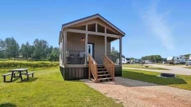 Una pequeña cabaña de madera con porche y escaleras, rodeada de hierba y árboles.