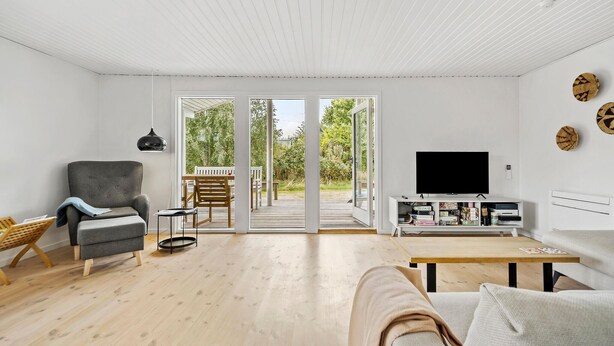 A modern living room with a grey sofa, a wooden coffee table, and a flat-screen TV on a white stand. There is a wooden deck visible through the glass doors leading outside. - Gorgeous home in Sjællands Odde with WiFi (Sjællands Odde)