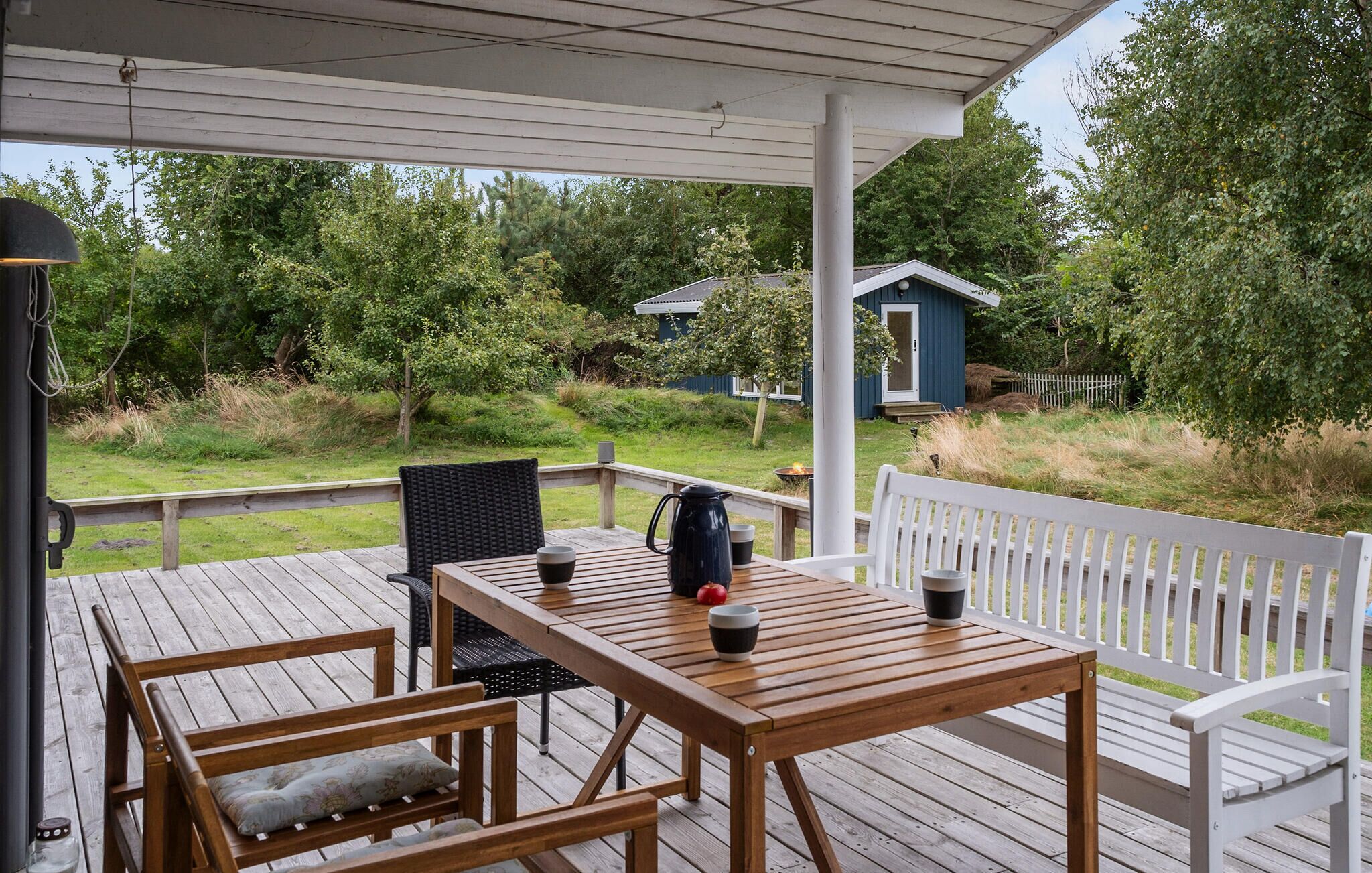A wooden deck with a table and chairs, a white bench, and a small blue building in the background.