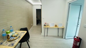 A hotel room with a wooden table, a black chair, a small table with items, a fire extinguisher, and a door.