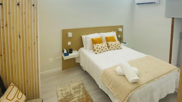A modern hotel room with a wooden headboard, a large bed with white bedding and decorative pillows, a bedside table with a lamp, and a small seating area with a chair and a patterned rug.